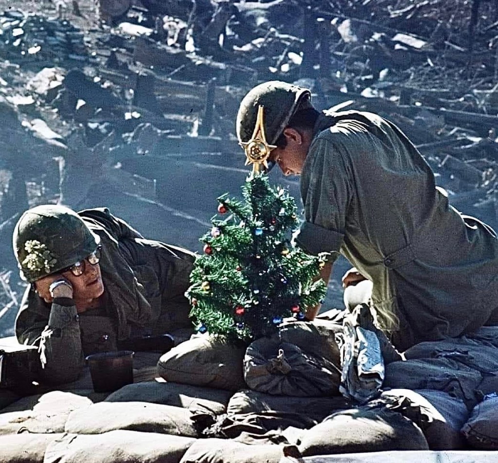 Two soldiers decorating a small Christmas tree among sandbags